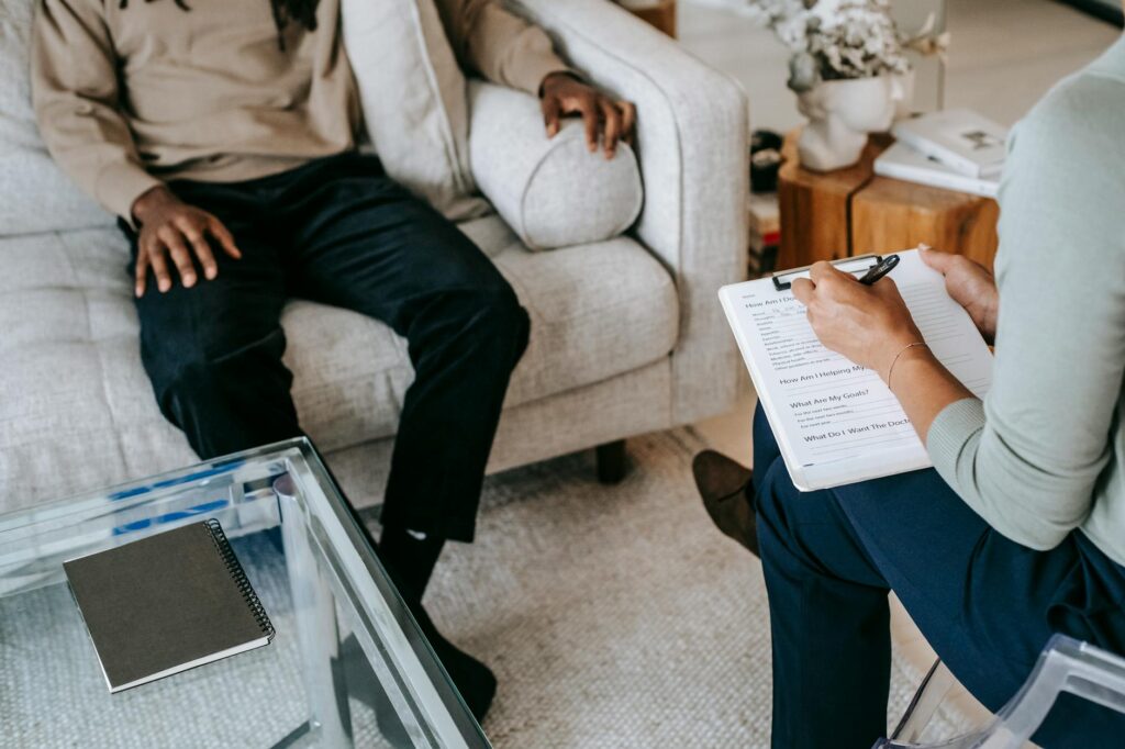 D'en haut, un homme afro-américain vêtu de vêtements décontractés est assis sur un canapé confortable et répond aux questions d'une consultante professionnelle dans un bureau de psychothérapie moderne