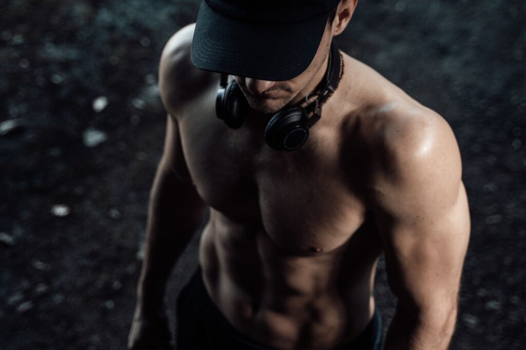 Homme torse nu portant des écouteurs, mettant en valeur sa forme physique et sa force dans un environnement sombre.