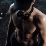 Homme torse nu portant des écouteurs, mettant en valeur sa forme physique et sa force dans un environnement sombre.